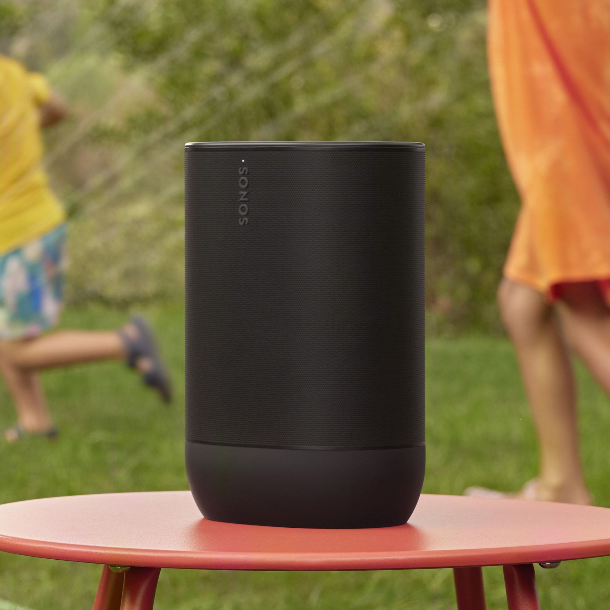 Close-up of a black cylindrical speaker on a pink table outdoors, with blurred people in colorful shirts in the background.
