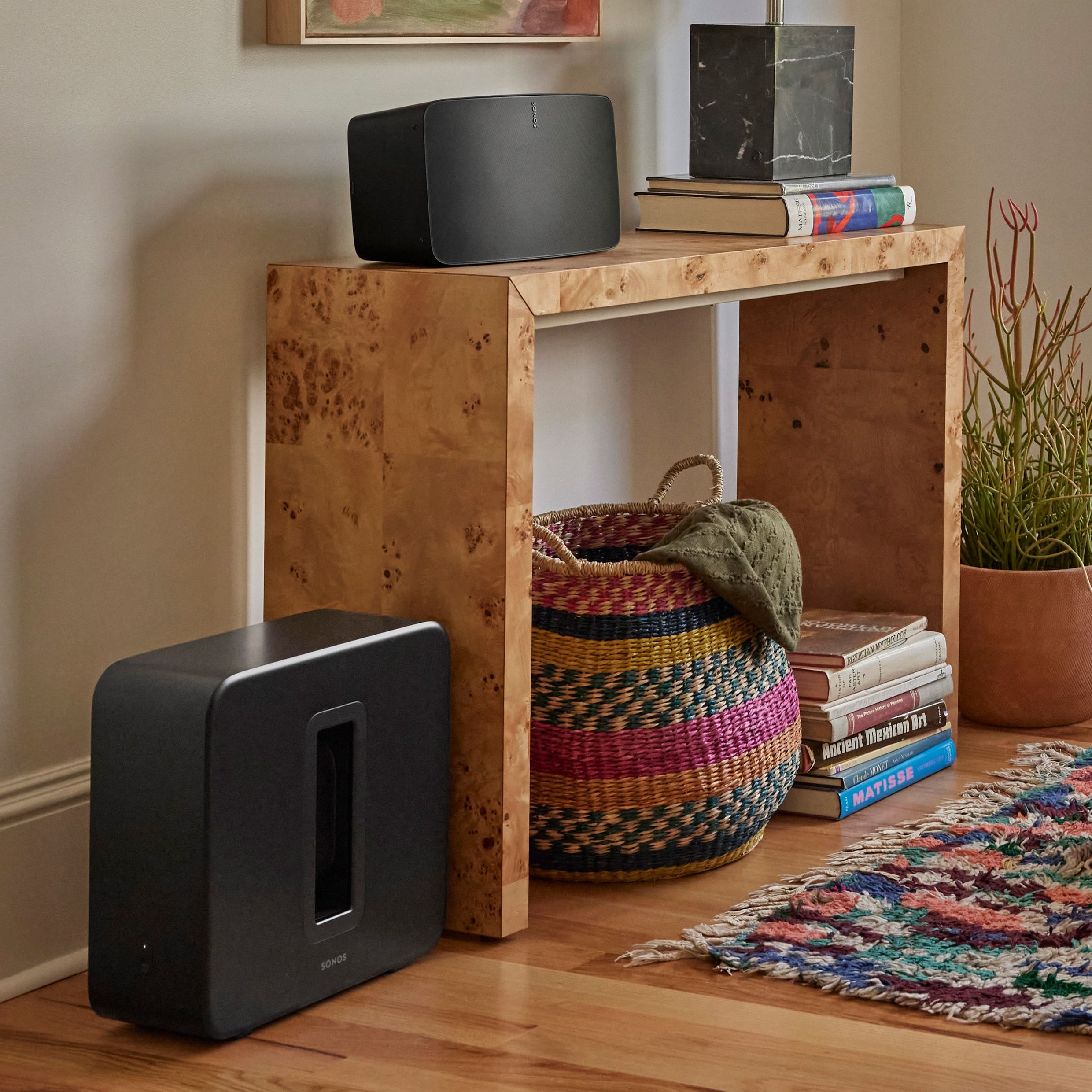 Black Sonos wireless speaker resting on a wooden console with books, baskets, and a woven rug nearby.