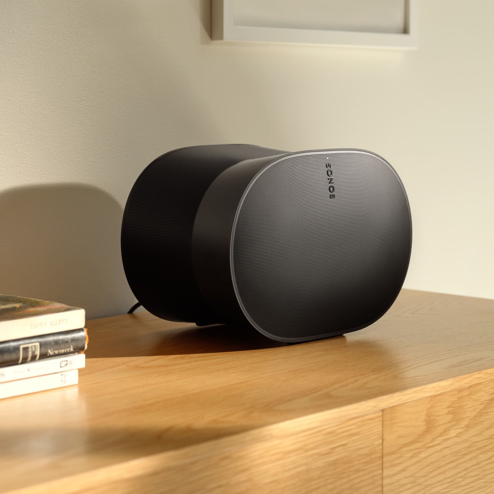 Black curved bookshelf speaker on a wooden desk with a small stack of books to the left.