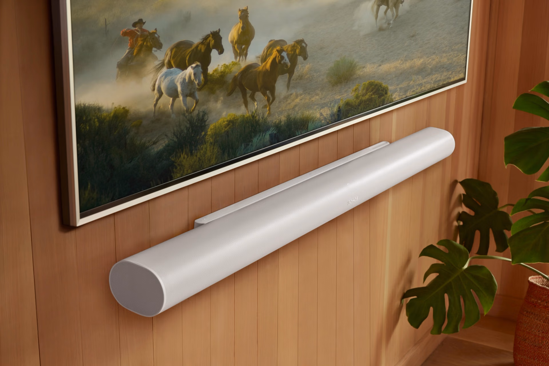 Wall-mounted TV shows a herd of running horses on a dusty plain; a white cylindrical soundbar sits below on a wood-paneled wall.