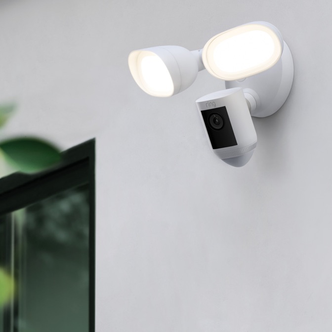 White outdoor security camera with two floodlights mounted on a wall, ring-brand lens facing outward at dusk.