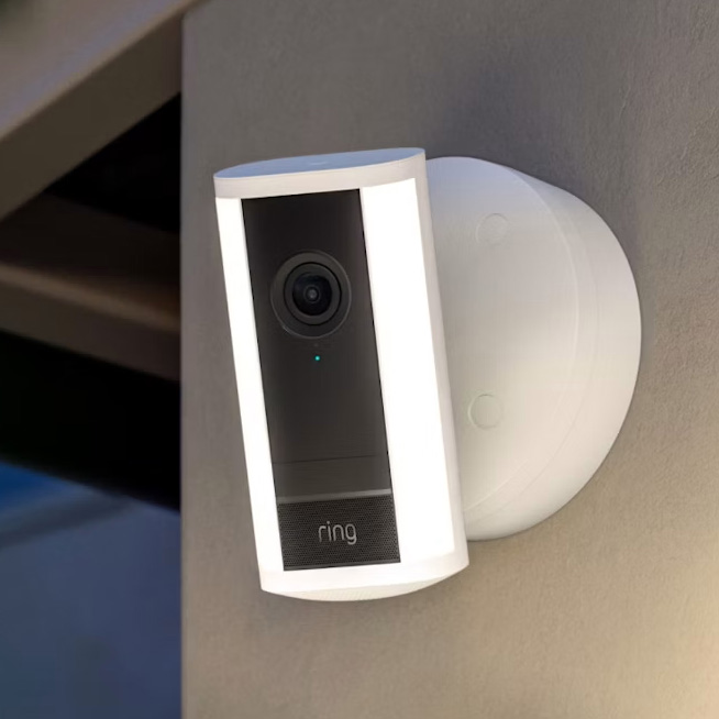 Ring doorbell camera mounted on a wall, with a white protective frame and circular mounting plate nearby, facing outward at dusk/dawn.