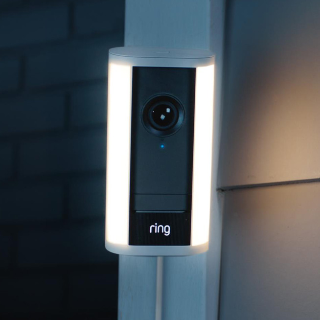 Ring video doorbell mounted on an exterior wall with vertical LED light strips glow on dusk/dawn. Description emphasizes the camera and lighting, indicating a security device exterior.